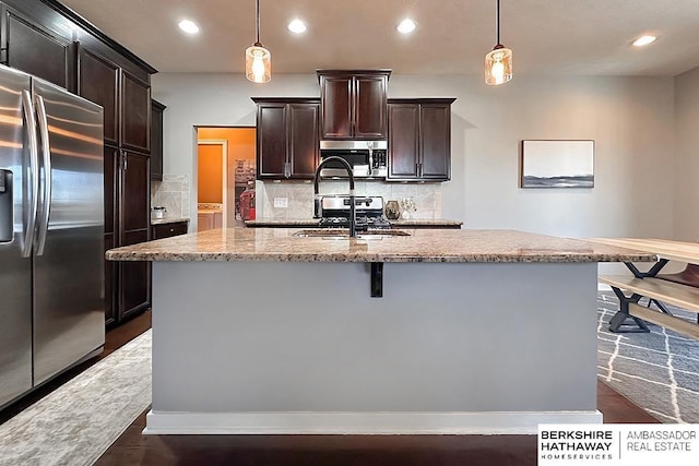 kitchen with light stone counters, a kitchen island with sink, a sink, appliances with stainless steel finishes, and backsplash