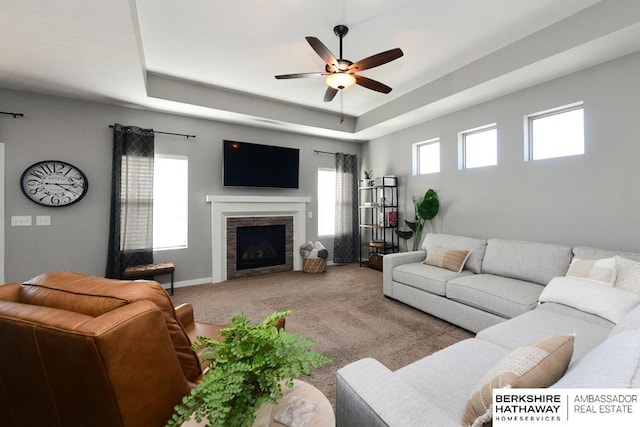 living room featuring a stone fireplace, a raised ceiling, carpet flooring, and a ceiling fan