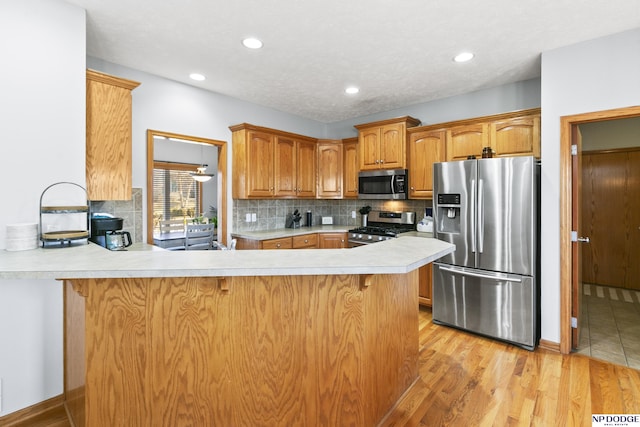 kitchen featuring light wood-type flooring, appliances with stainless steel finishes, a peninsula, light countertops, and decorative backsplash