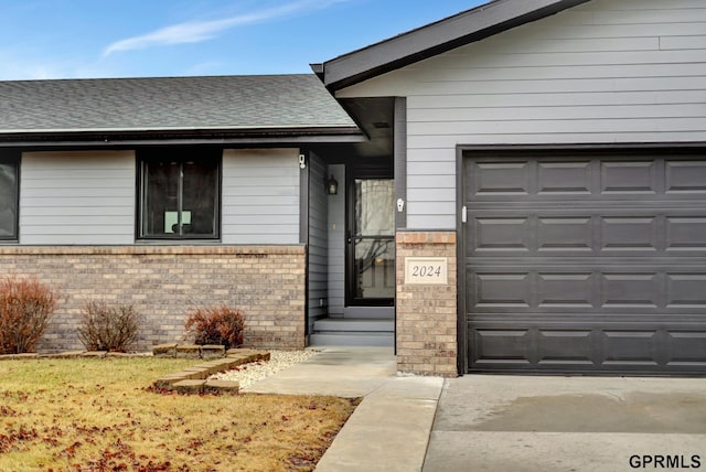 entrance to property featuring an attached garage, brick siding, and roof with shingles