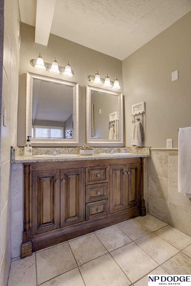 bathroom with tile walls, double vanity, wainscoting, a textured ceiling, and a sink