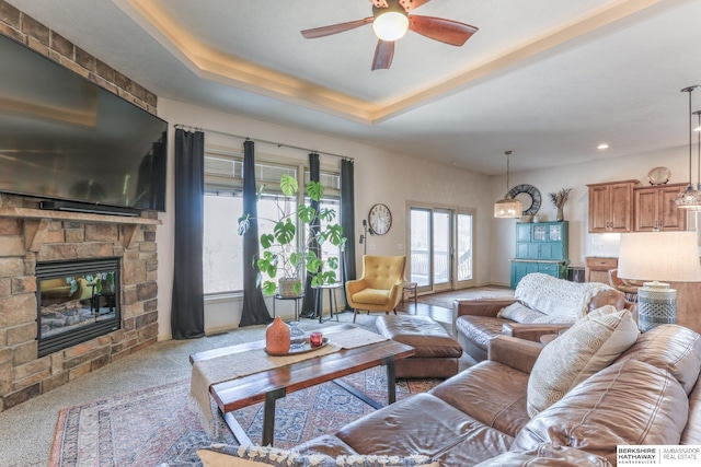 carpeted living area with a stone fireplace, recessed lighting, a raised ceiling, and a ceiling fan