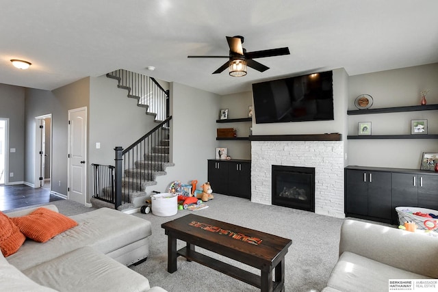 living area featuring baseboards, a ceiling fan, a glass covered fireplace, and stairs