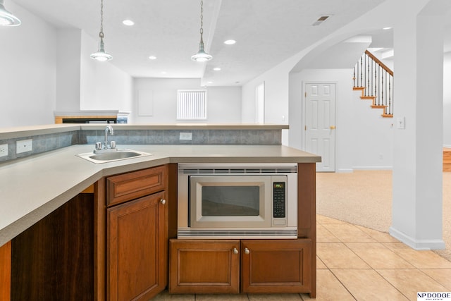 kitchen with a sink, stainless steel microwave, decorative light fixtures, brown cabinetry, and light colored carpet