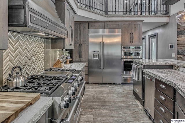 kitchen featuring dark brown cabinetry, light stone countertops, stainless steel appliances, and exhaust hood