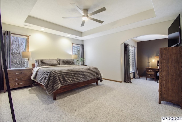 bedroom with light colored carpet, a raised ceiling, arched walkways, and baseboards