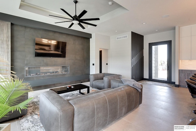 living room with visible vents, ceiling fan, a glass covered fireplace, tile walls, and a raised ceiling