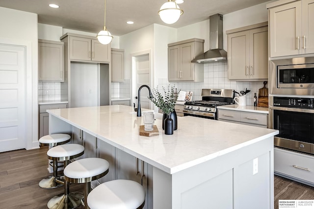 kitchen featuring a center island with sink, dark wood-style flooring, gray cabinetry, appliances with stainless steel finishes, and wall chimney range hood