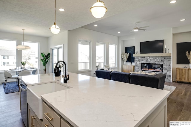 kitchen featuring open floor plan, light stone counters, a stone fireplace, a ceiling fan, and dark wood-style flooring