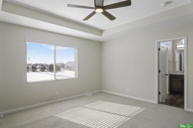 unfurnished bedroom featuring a raised ceiling, a ceiling fan, ensuite bath, carpet floors, and baseboards