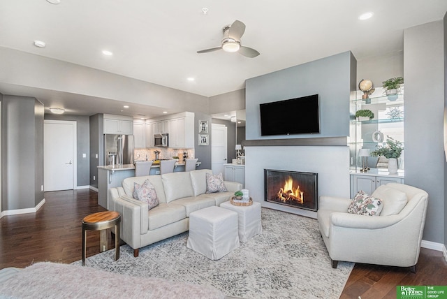 living room with dark wood finished floors, a glass covered fireplace, baseboards, and a ceiling fan