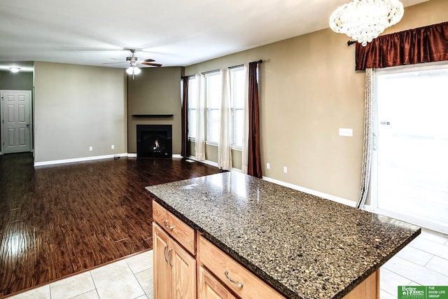 kitchen featuring dark stone countertops, baseboards, light tile patterned flooring, a fireplace, and ceiling fan with notable chandelier