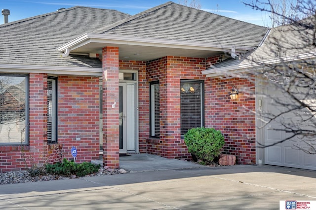 entrance to property with driveway, an attached garage, brick siding, and roof with shingles