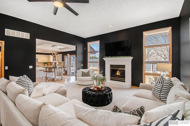 living area with a tray ceiling, a brick fireplace, ceiling fan with notable chandelier, and visible vents