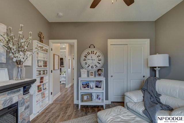 living area with baseboards, a stone fireplace, wood finished floors, and a ceiling fan