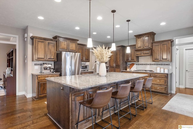 kitchen featuring a sink, stainless steel appliances, a center island with sink, dark wood-type flooring, and light stone counters