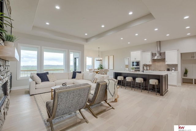 living room with a stone fireplace, light wood-type flooring, recessed lighting, a chandelier, and a tray ceiling