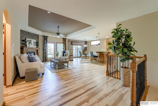 living area featuring light wood-style flooring, a raised ceiling, a tiled fireplace, ceiling fan, and baseboards