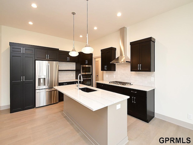 kitchen with a kitchen island with sink, appliances with stainless steel finishes, a sink, light wood-style floors, and wall chimney range hood