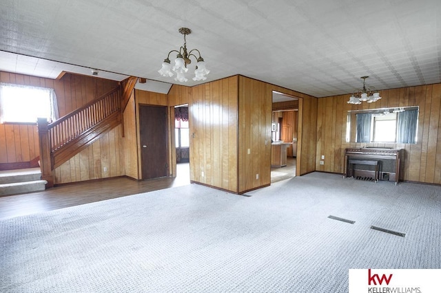 unfurnished living room featuring wooden walls, carpet flooring, a notable chandelier, and stairway