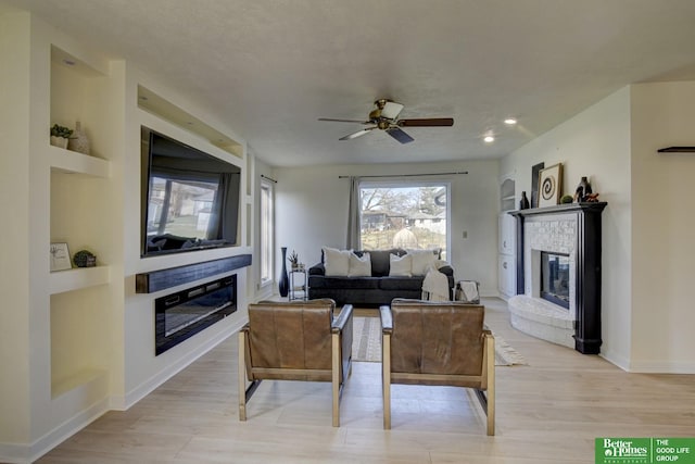 living area featuring a ceiling fan, a glass covered fireplace, light wood-style flooring, and built in shelves