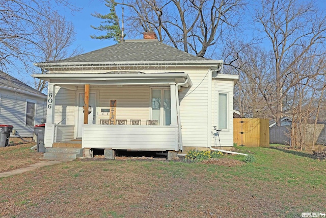 view of front facade featuring a front yard, covered porch, a chimney, fence, and a shingled roof