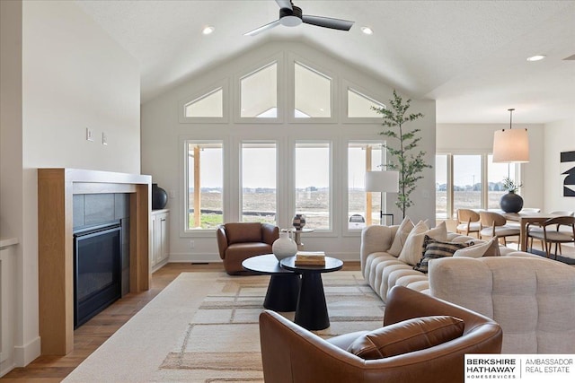living area with ceiling fan, light wood-type flooring, a tile fireplace, baseboards, and lofted ceiling