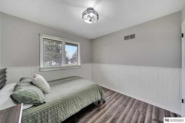 bedroom with a textured ceiling, visible vents, wainscoting, and wood finished floors