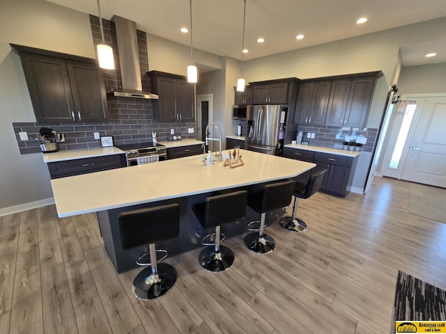 kitchen featuring wall chimney exhaust hood, a kitchen bar, light wood-type flooring, light countertops, and stainless steel appliances
