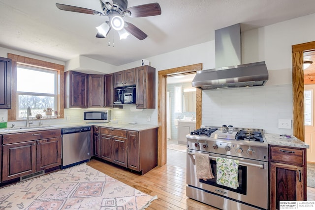 kitchen featuring wall chimney exhaust hood, light wood-style flooring, a ceiling fan, appliances with stainless steel finishes, and a sink