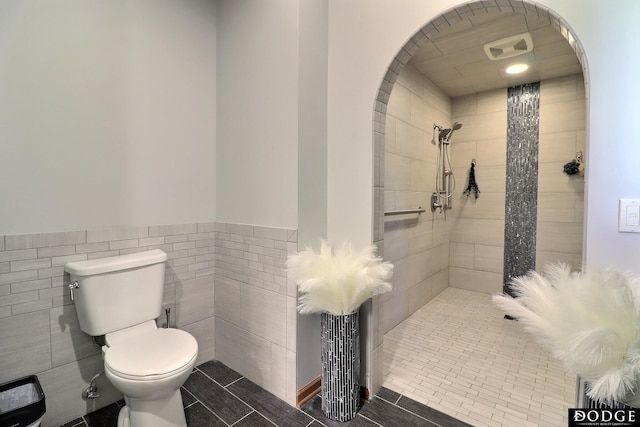 bathroom with tiled shower, tile patterned flooring, a wainscoted wall, toilet, and tile walls