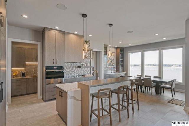 kitchen featuring stainless steel oven, a center island with sink, modern cabinets, a healthy amount of sunlight, and decorative backsplash