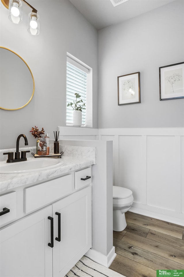 half bathroom featuring a decorative wall, vanity, a wainscoted wall, toilet, and wood finished floors