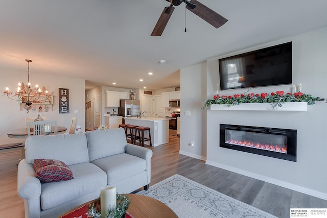 living area featuring a glass covered fireplace, baseboards, recessed lighting, light wood finished floors, and ceiling fan with notable chandelier
