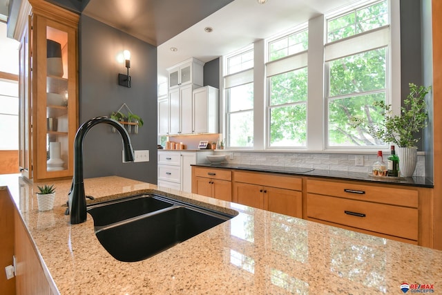 kitchen with black electric cooktop, a sink, glass insert cabinets, light stone countertops, and tasteful backsplash