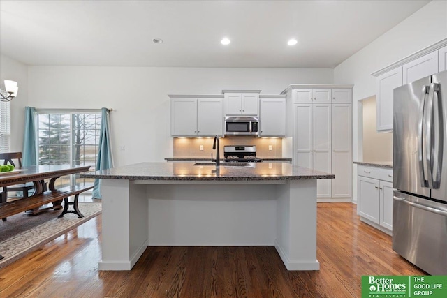kitchen with stainless steel appliances, a sink, dark stone countertops, a center island with sink, and wood finished floors