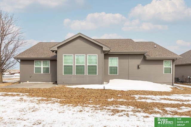 snow covered house with cooling unit, a patio area, and roof with shingles