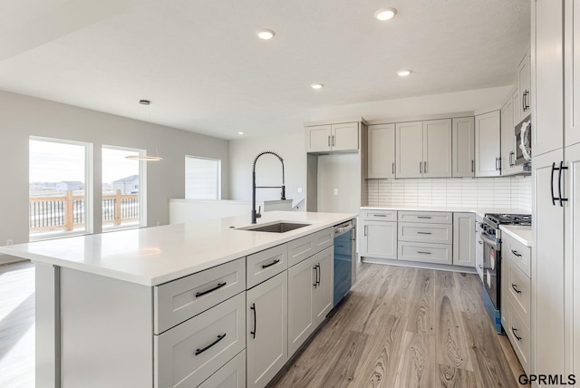 kitchen featuring a sink, an island with sink, light wood-type flooring, stainless steel appliances, and light countertops