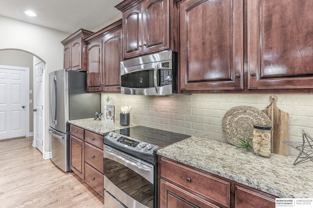 kitchen featuring decorative backsplash, arched walkways, light stone counters, light wood finished floors, and stainless steel appliances