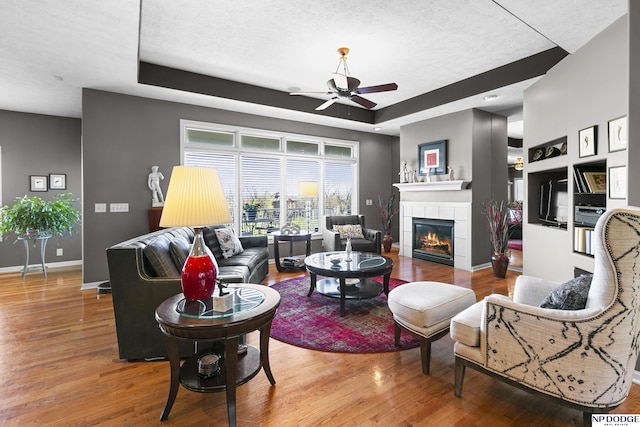 living room with a ceiling fan, wood finished floors, a tray ceiling, a tiled fireplace, and baseboards