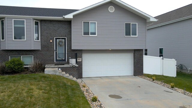 raised ranch with a front yard, a garage, brick siding, fence, and concrete driveway