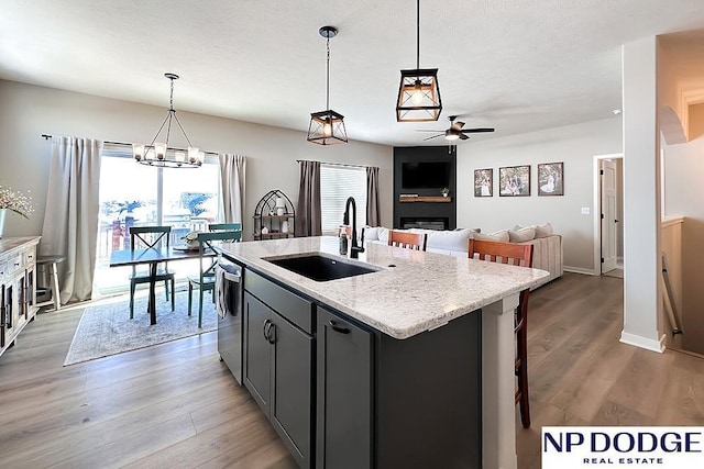 kitchen featuring stainless steel dishwasher, a sink, light wood-type flooring, a center island with sink, and ceiling fan with notable chandelier
