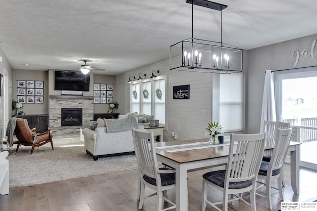 dining area featuring a stone fireplace, a textured ceiling, light wood-style flooring, and ceiling fan with notable chandelier