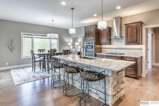 kitchen with wall chimney range hood, a sink, a breakfast bar area, appliances with stainless steel finishes, and light stone countertops