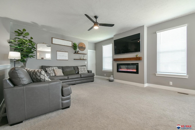 carpeted living area with ceiling fan, a textured ceiling, a glass covered fireplace, baseboards, and visible vents