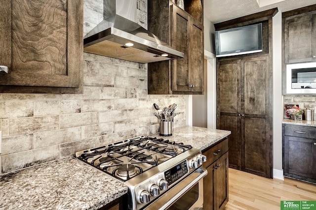kitchen featuring wall chimney range hood, light wood-style floors, light stone countertops, stainless steel gas range, and dark brown cabinetry