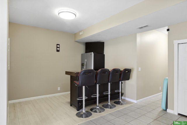 bar featuring light wood-type flooring, stainless steel fridge with ice dispenser, visible vents, and baseboards