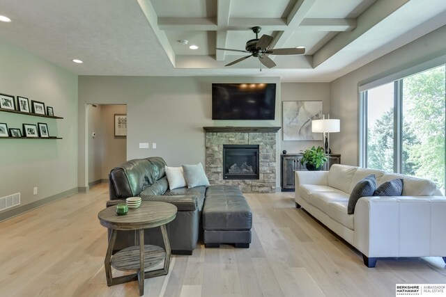 living room featuring ceiling fan, beam ceiling, light wood finished floors, a stone fireplace, and coffered ceiling