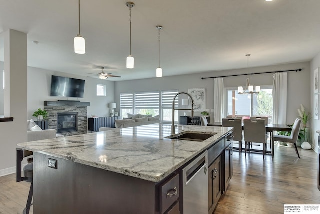 kitchen featuring a stone fireplace, stainless steel dishwasher, a center island with sink, a sink, and light wood-style floors