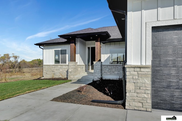 view of exterior entry with board and batten siding, stone siding, roof with shingles, a lawn, and a garage